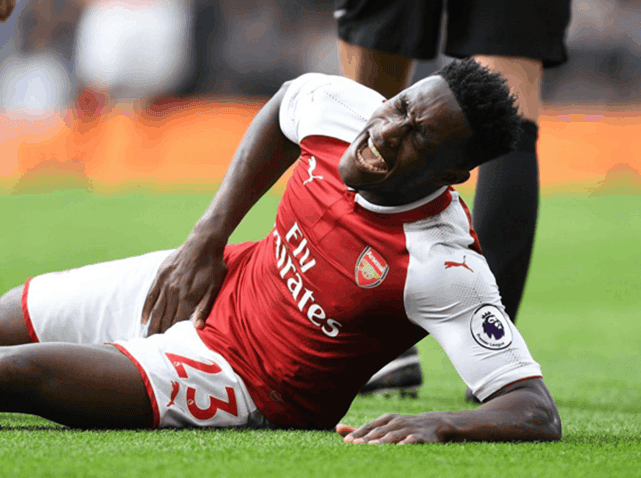 A footballer in a red and white kit lying on the pitch, grimacing and holding his hip