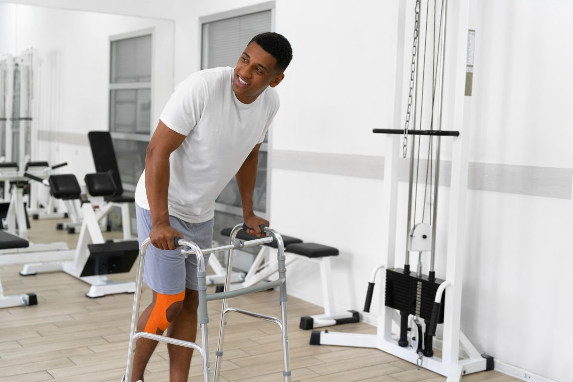 A patient with a bandaged knee walking with a support frame in a rehabilitation gym.