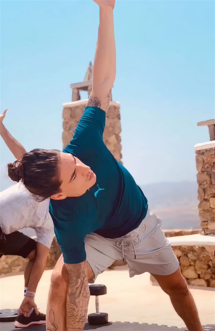 A person practising a standing side-bend yoga posture outdoors in bright sunlight.