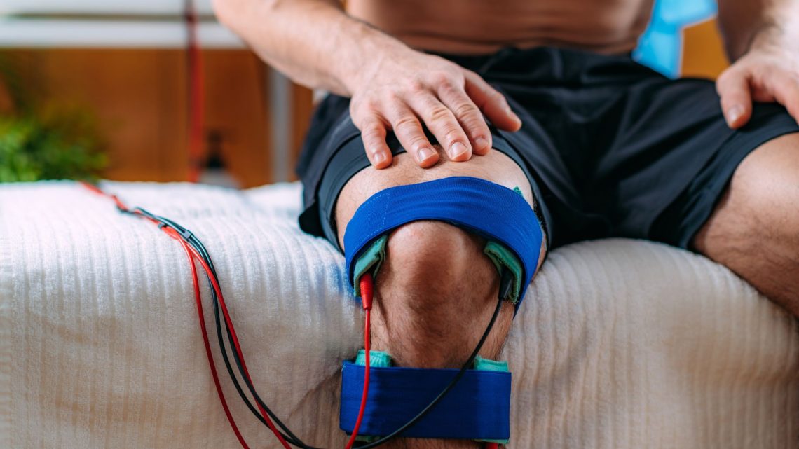 An electrode wrap fitted around a patient's knee with leads connected to an electrotherapy unit.