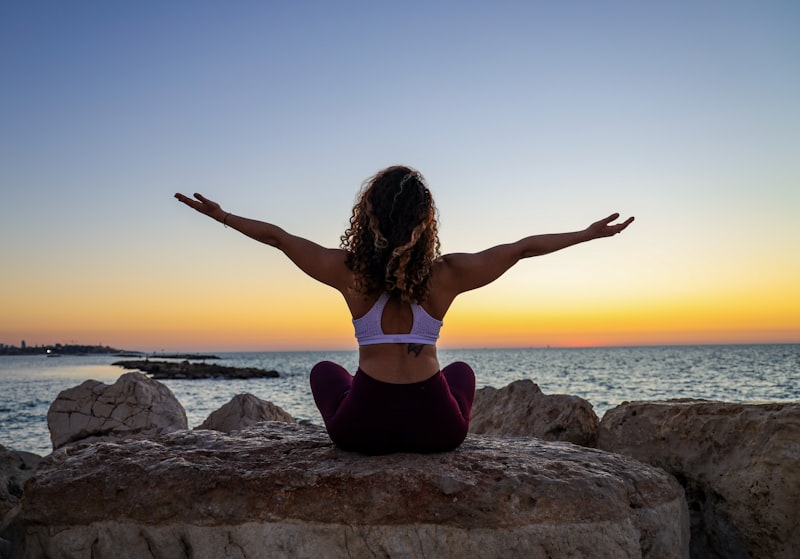 Woman sitting cross-legged on a rock by the sea at sunrise with her arms outstretched